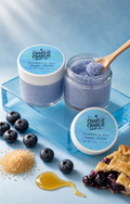 Two jars of Charlie Charlie Soap Co. Blueberry Pie Sugar Scrub are displayed on a light blue surface with fresh blueberries, brown sugar, honey, and a blueberry pie crust.