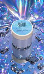 Charlie Charlie Soap Co. Blueberry Pie Sugar Scrub in a glittery silver jar with a light blue and white lid, styled on a reflective blue surface surrounded by holographic bubbles, stars, and chrome soap shapes.
