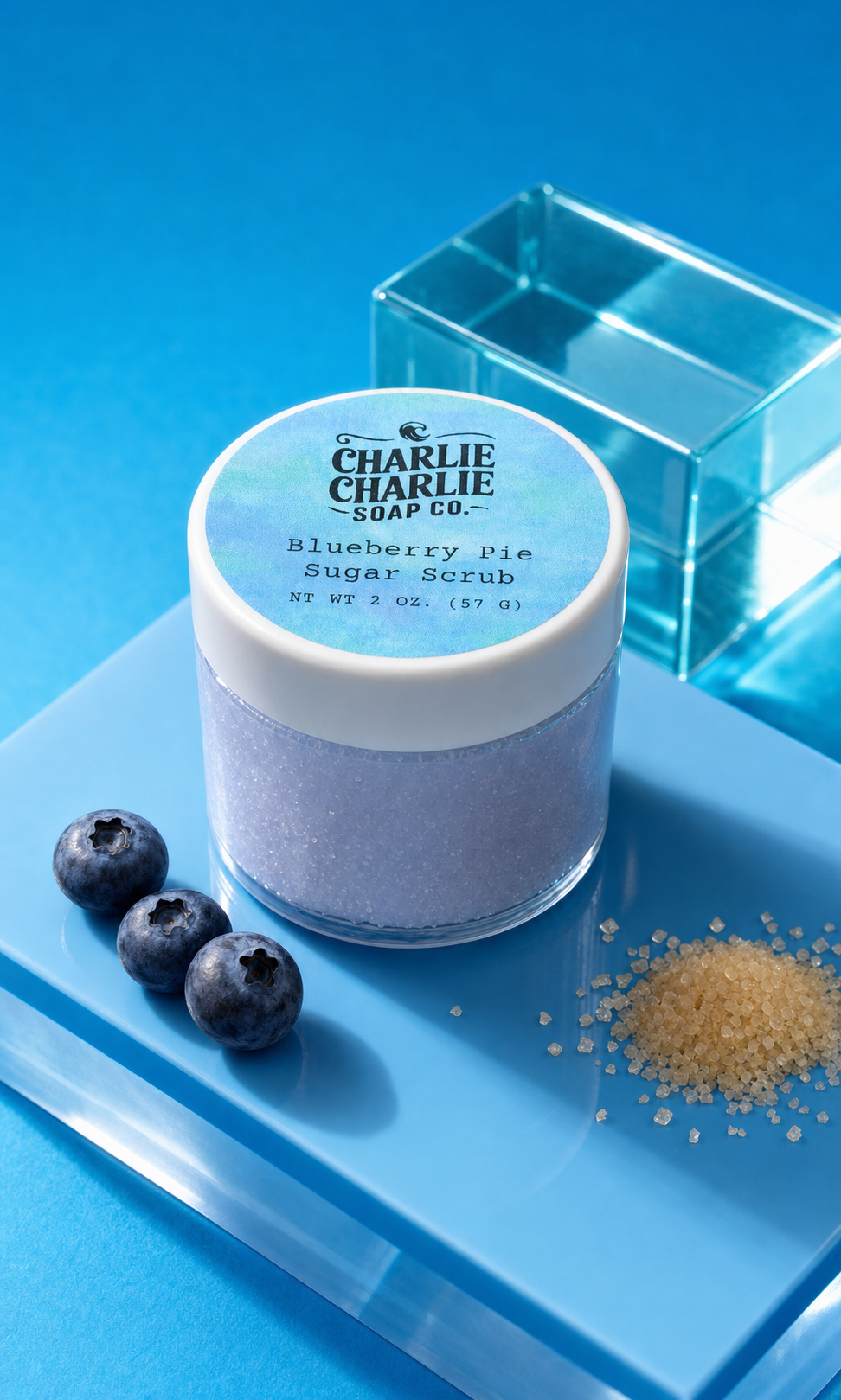 A jar of Charlie Charlie Soap Co. Blueberry Pie Sugar Scrub sits on a blue tray with three blueberries and a small pile of sugar crystals, with a clear acrylic block in the background.