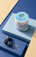 A small jar of Charlie Charlie Soap Co. Blueberry Pie Sugar Scrub sits on a light blue pedestal, with three blueberries and scattered sugar crystals on a dark blue surface beside it.