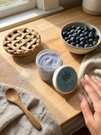A jar of blueberry pie scrub by Charlie Charlie Soaps sits open on a wooden table next to a blueberry pie, a bowl of fresh blueberries, and a hand reaching toward the lid. The jar has a light blue label with the brand name visible.
