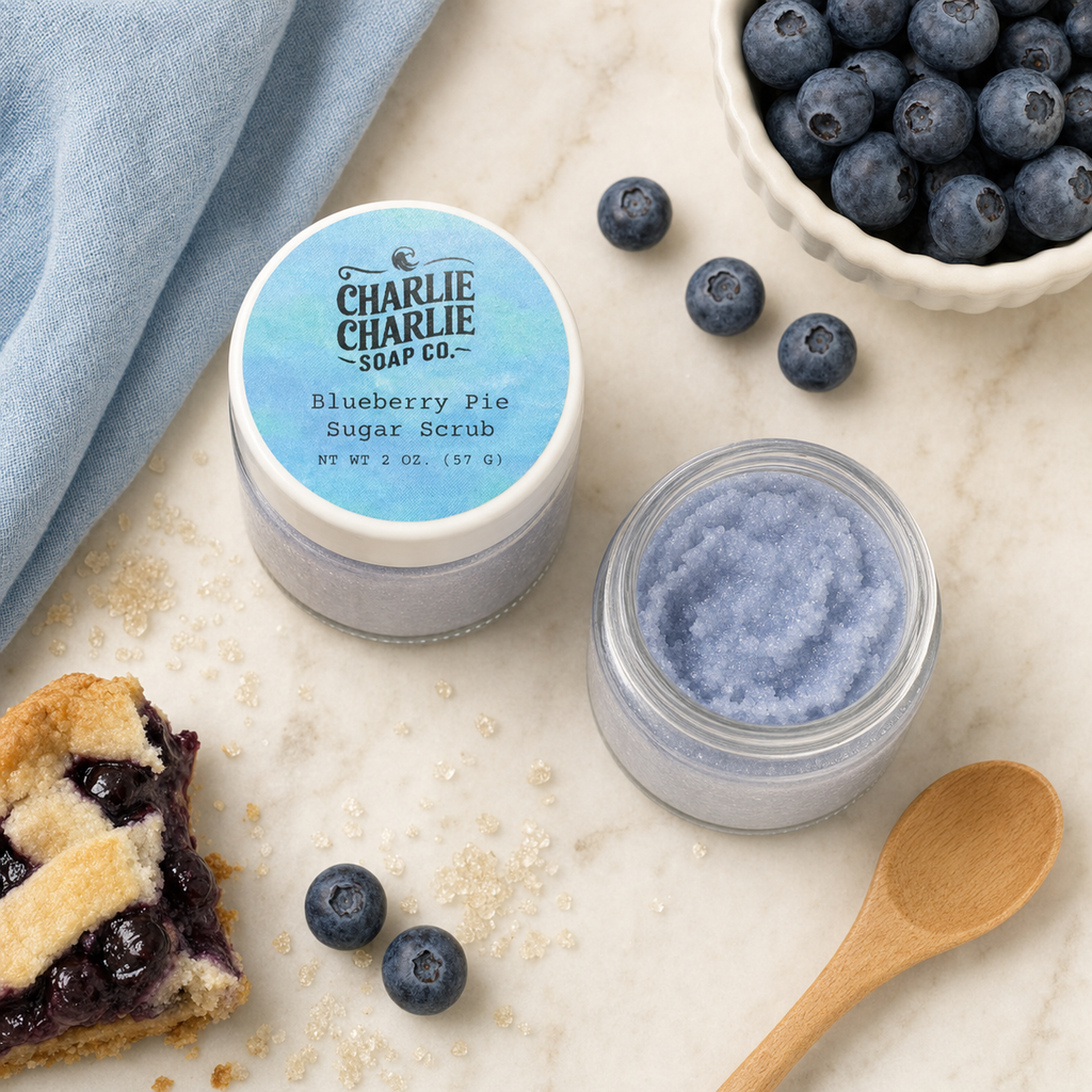 An open jar of blueberry pie sugar scrub by Charlie Charlie Soap Co. sits on a marble surface next to an unopened jar with a blue label, styled with blueberries, a pie slice, a wooden spoon, and a blue cloth.