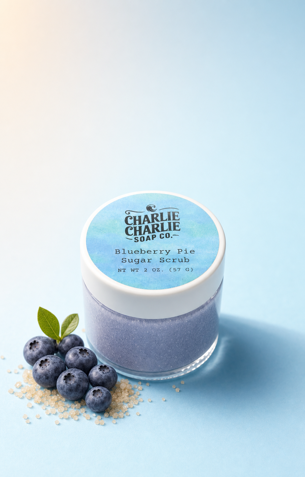 A jar of Charlie Charlie Soap Co. Blueberry Pie Sugar Scrub, featuring a white lid with a blue watercolor-style label, set next to fresh blueberries and sugar crystals on a light blue background.