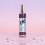 A bottle of Charlie Charlie Soap Co. Classic Starfruit Body Oil with purple glitter liquid inside, set on a clear acrylic stand with scattered decorative crystals against a pink and purple gradient background.