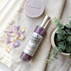Charlie Charlie Soap Co. Stardust Chemicall Stimulant Body Oil in a clear bottle with a silver cap, styled on a beige linen cloth with dried lavender petals, a round soap bar, and a small potted eucalyptus plant.