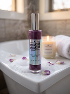 Charlie Charlie Soap Co. Cosmic Stardust Body Oil in a clear bottle with a silver cap, set in a bathroom scene on a white tub with bubbles, rose petals, a lit candle, and a towel.