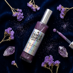 A bottle of Charlie Charlie Soap Co. Cosmic Stardust Body Oil surrounded by purple dried flowers, crystals, and a dropper on a dark navy velvet background.