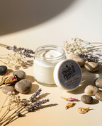 Jar of cream with a lid labeled 'Charlie Charlie Soap Co.' on a beige surface with stones and lavender flowers.