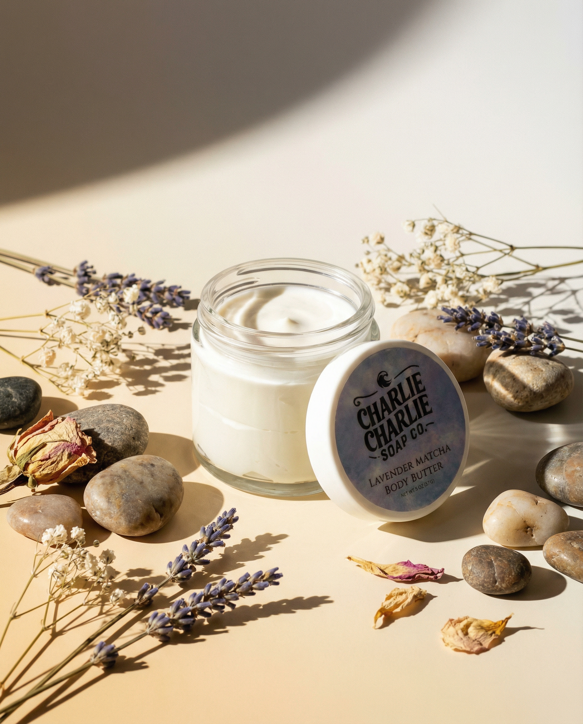 Jar of cream with a lid labeled 'Charlie Charlie Soap Co.' on a beige surface with stones and lavender flowers.