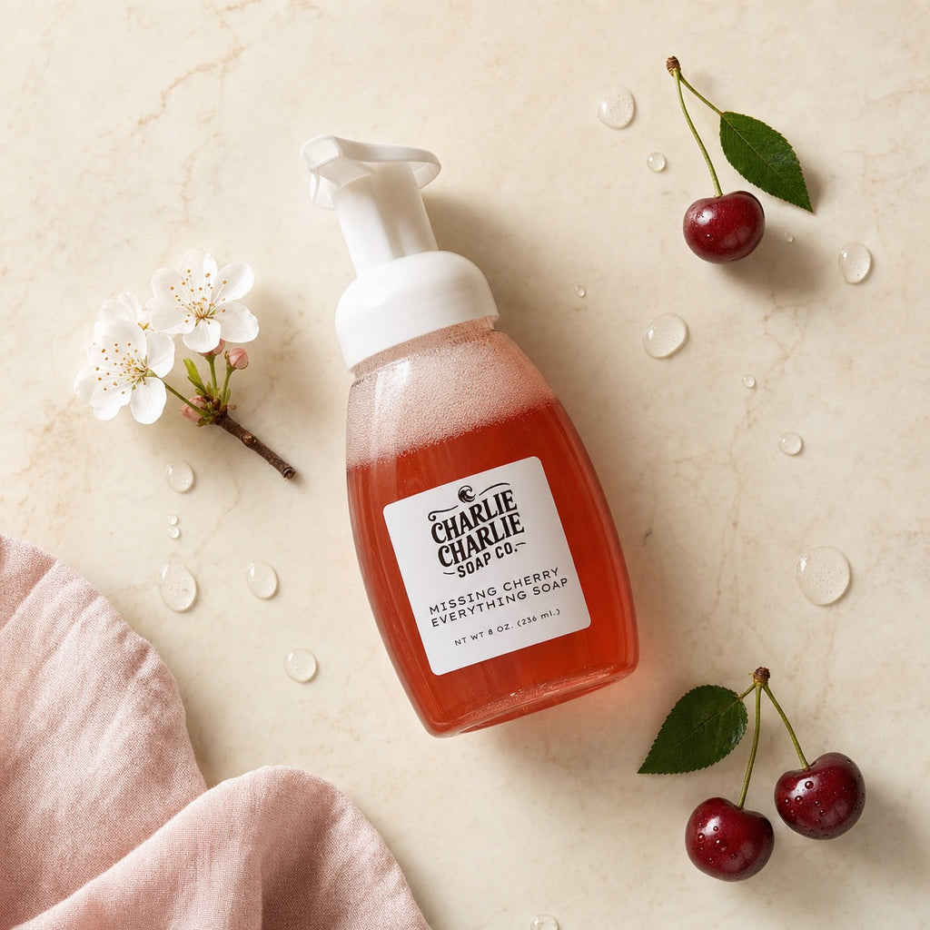 Charlie Charlie Soap Co. Missing Cherry Everything Fresh soap in a frosted bottle with a white pump, styled on a beige marble surface with cherry props, white blossoms, water droplets, and a soft pink cloth.