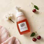 Charlie Charlie Soap Co. Missing Cherry Everything Fresh soap in a frosted bottle with a white pump, styled on a beige marble surface with cherry props, white blossoms, water droplets, and a soft pink cloth.