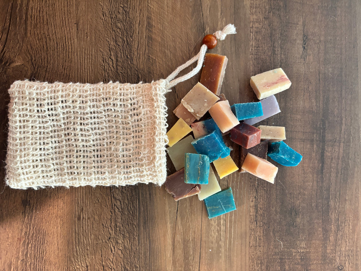 A natural beige cotton muslin soap bag with a drawstring and wooden bead, shown on a dark wood background with colorful handmade soap bars spilling out.