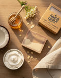 Bar of soap, jar of honey, coconut, and bowl of cream on a wooden surface with 'Charlie Chaplin Soap Co.' packaging.