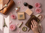 Collection of beauty and travel items on a pink surface with flowers and a hand holding a book.