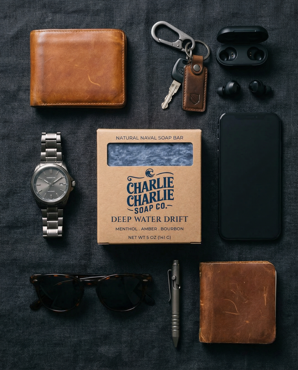 Collection of everyday carry items including a wallet, watch, phone, sunglasses, pen, and soap on a dark surface.