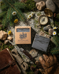 Bar of soap from Charlie Charlie Soap Co. on a natural setting with compass, leather pouch, and camping gear.