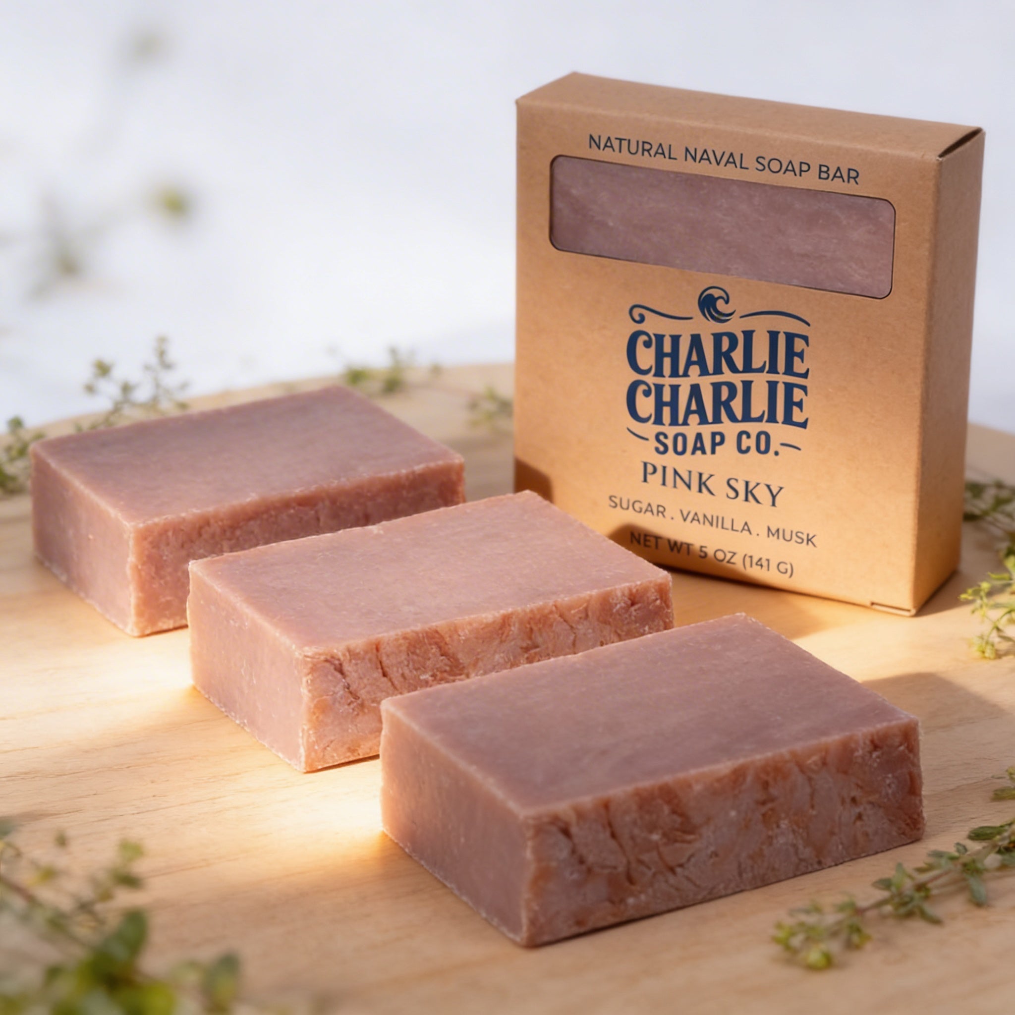Three bars of pink soap with a cardboard box labeled 'Charlie Charlie Soap Co. Pink Sky' on a wooden surface.