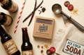 Root beer bottles, vanilla beans, and a box of Charlie Charlie soap on a light background