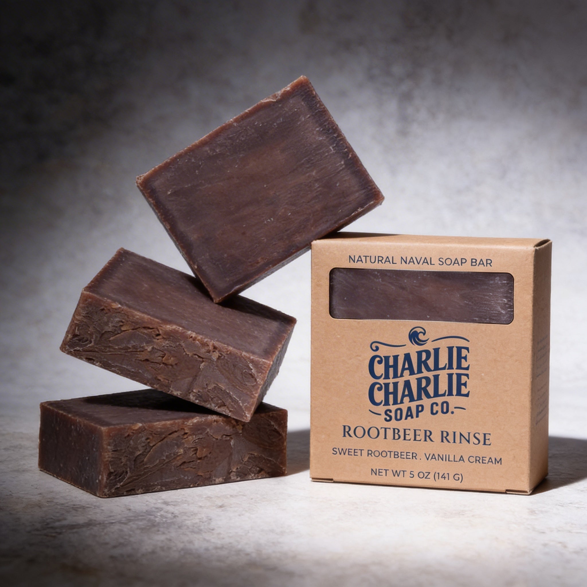 Three bars of Charlie Charlie Soap Co. root beer rinse soap with their packaging on a gray background.