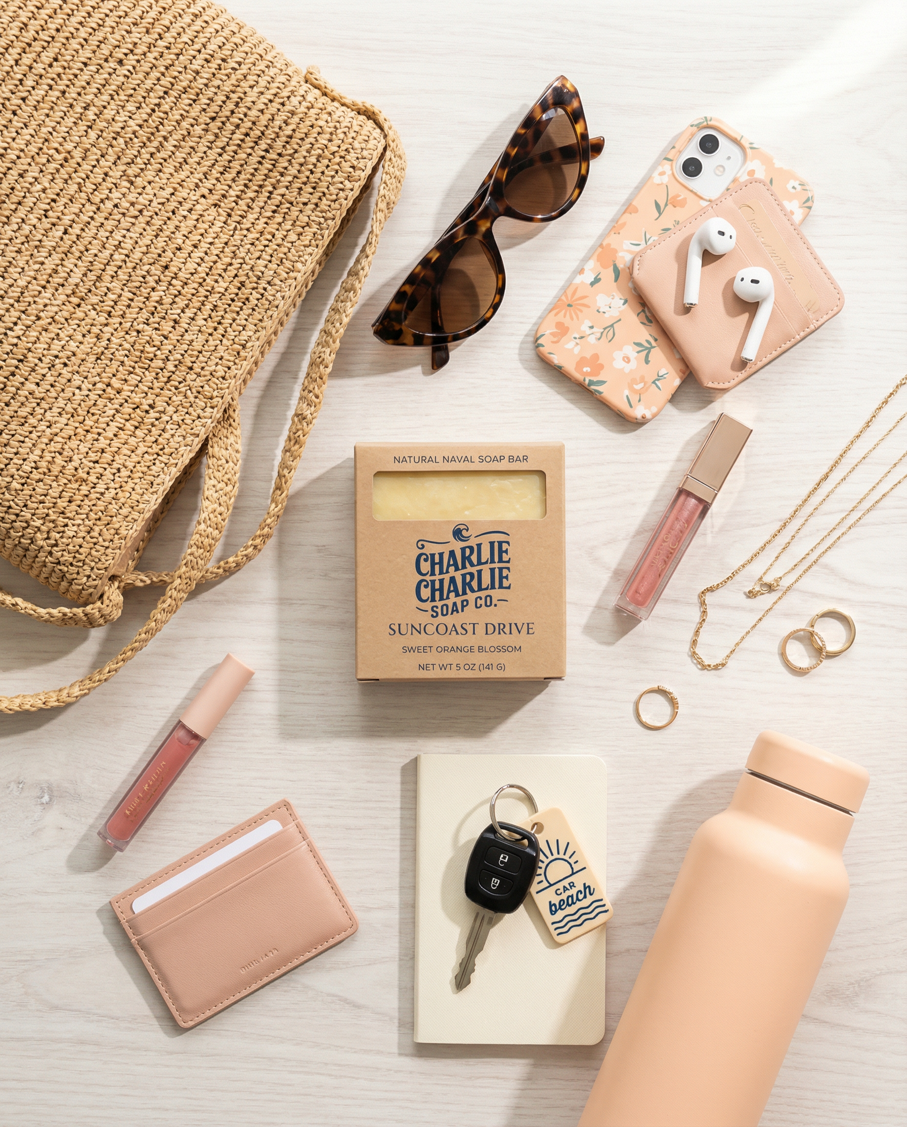 Collection of everyday items including a straw bag, sunglasses, phone case, lip gloss, soap, keychain, and water bottle on a light surface.